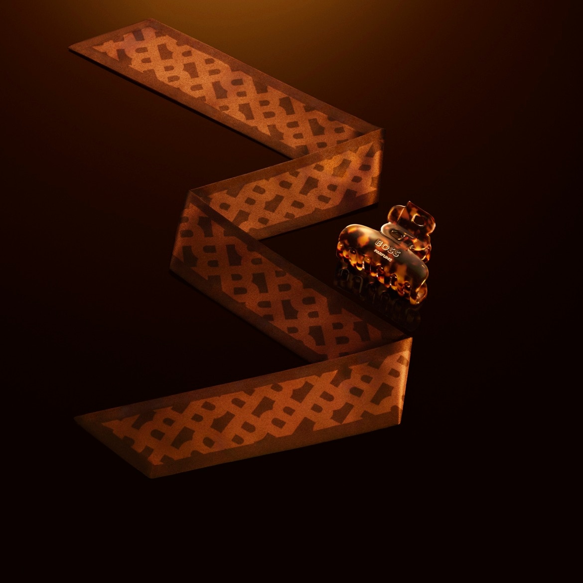 E braune Bändeli mit Muster und e Haarspange vo BOSS Parfums ligged ufem dunkle Untergrund.