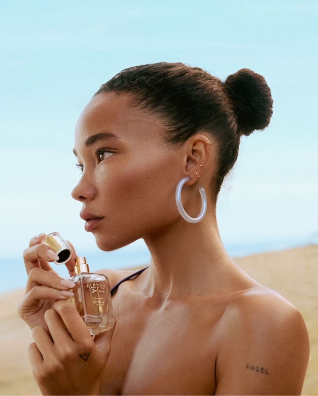 Une femme aux yeux bleus, les mains derrière la tête, porte une robe rose sur fond de mer calme.