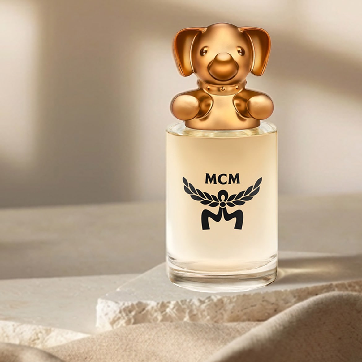 Flacon de parfum MCM en verre transparent avec un bouchon doré en forme de chien, le logo MCM noir est imprimé sur le devant.