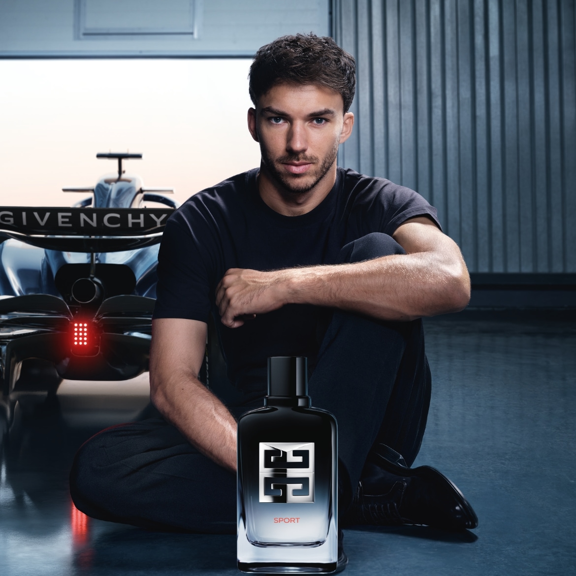 Ein Mann sitzt vor einem Rennwagen, neben ihm Givenchy Sport Parfüm, der Flakon ist schwarz mit silbernem Logo.