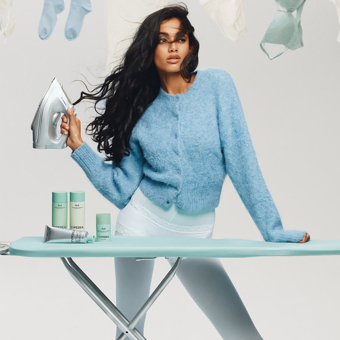 D'Frau hält en Eisen an der Hand, steet bei engem Bügelbrett mat TYPEBEA Hautpfleegprodukter, déi an hellgréng Behälter verpaakt sinn.