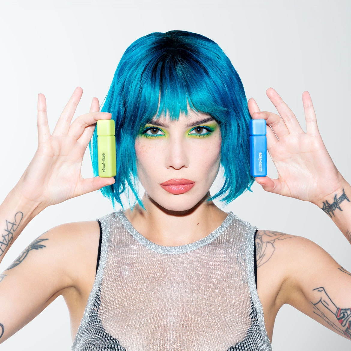 Une femme aux cheveux bleus présente deux produits : un flacon vert clair et un flacon bleu, pour un look coloré et tendance.
