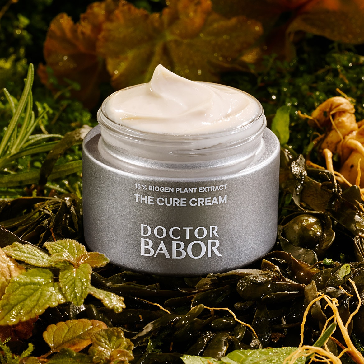 Die Doctor Babor The Cure Cream mit 15% Biogen Pflanzenextrakt in einem silbernen Tiegel mit einer Textur aus weißer Creme, die auf einem Bett aus Blättern und Pflanzen liegt.
