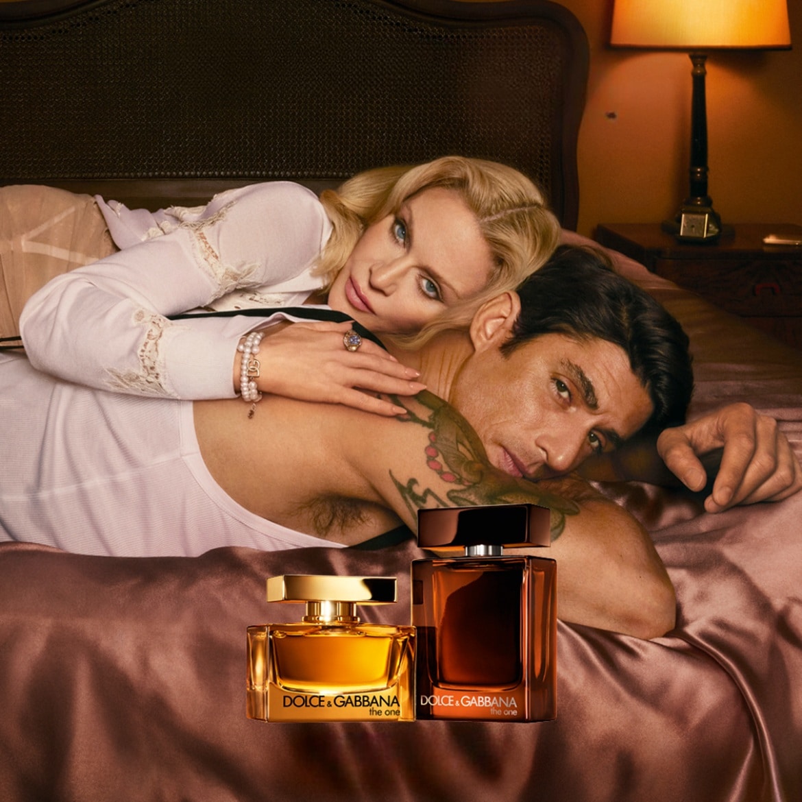 Sur un lit satiné, un couple enlacé présente les parfums "The One" de Dolce & Gabbana : elle, blond platine en chemise blanche, lui, torse nu avec un tatouage.