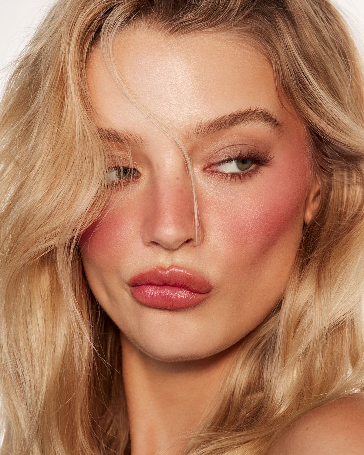Eine junge Frau mit blonden Haaren, die ihr Gesicht umrahmen, trägt ein natürliches Make-up mit rosa Rouge auf den Wangen und glänzenden Lippen.