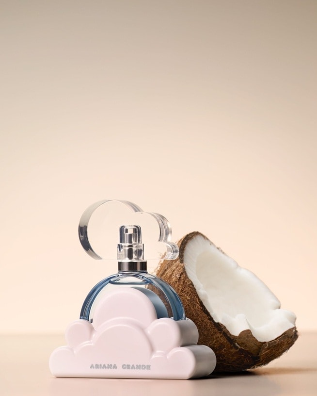 Un flacon de parfum en forme de nuage est placé à côté d’une noix de coco ouverte sur un fond clair. Cette association évoque des notes parfumées tropicales et fruitées ainsi que des créations olfactives aux accents estivaux.