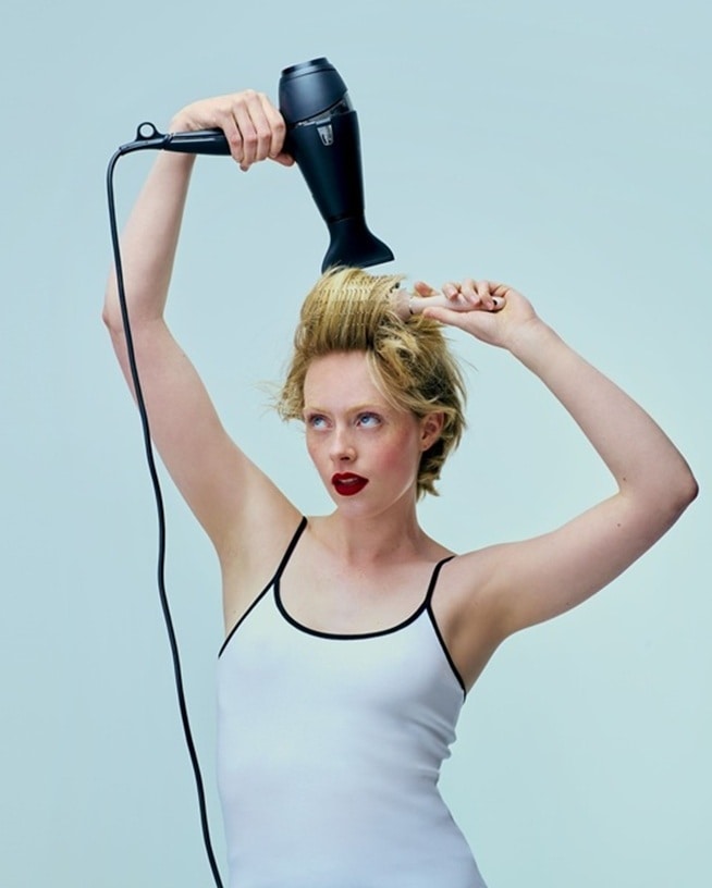 Une femme se sèche les cheveux blonds courts avec un sèche-cheveux noir et une brosse ronde, coiffant ses cheveux sans les abîmer grâce à nos hairstylers innovants.