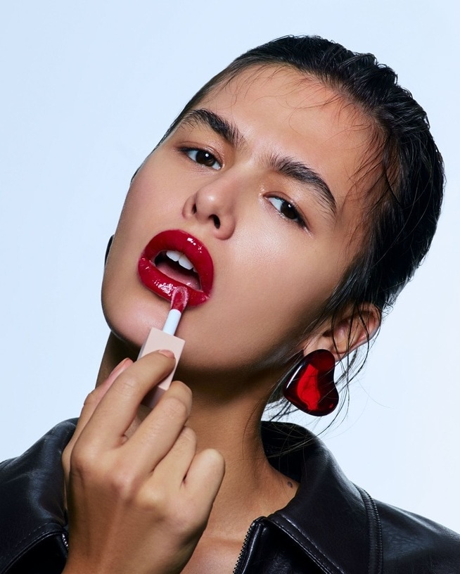 Eine Frau trägt einen glänzenden roten Lippenstift mit einem Applikator auf, dazu passende herzförmige Ohrringe und eine schwarze Lederjacke.'