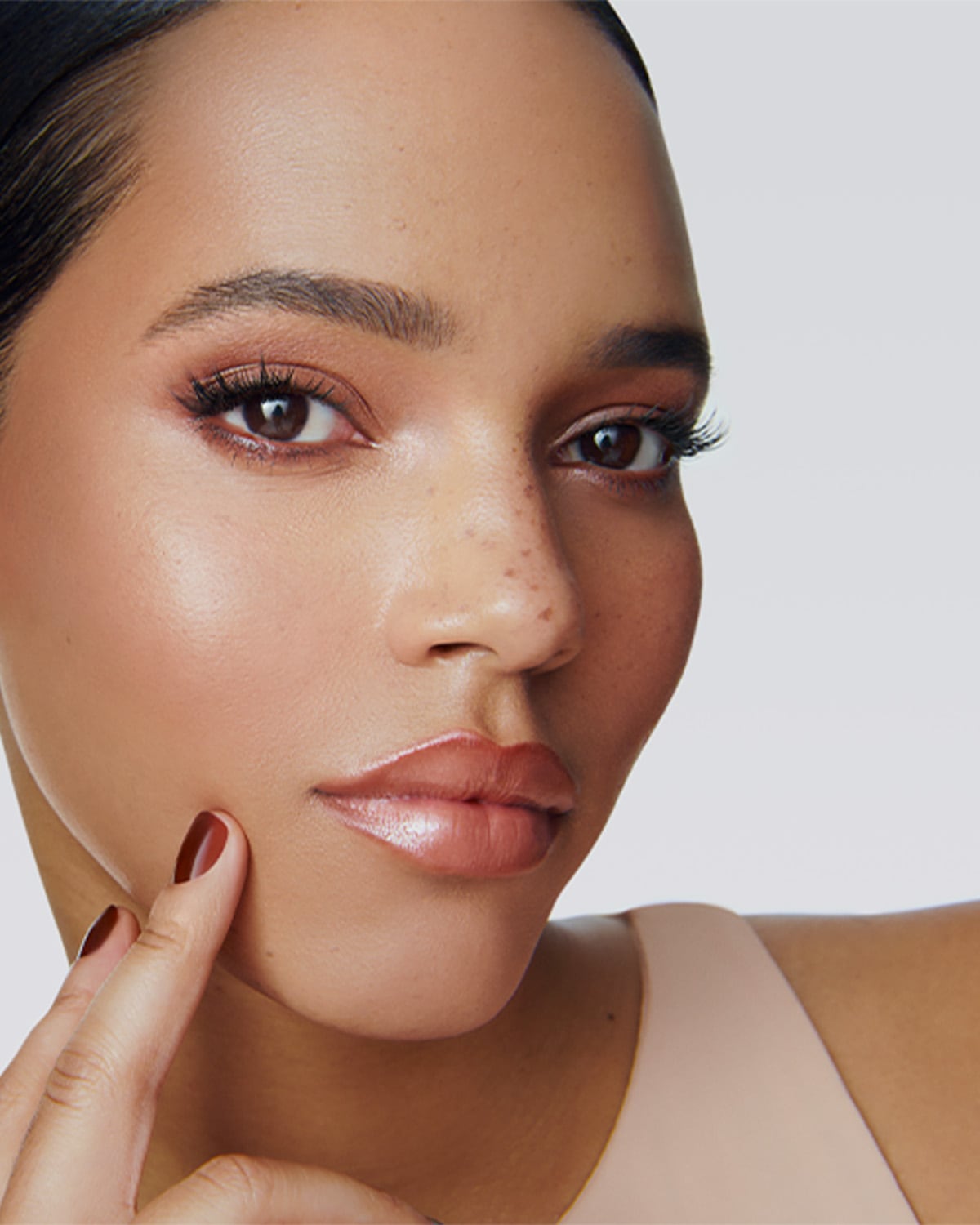 Nahaufnahme eines Frauengesichts mit braunen Augen, rosafarbenem Lidschatten und glänzenden Lippen, die mit einem Finger die Wange berührt.