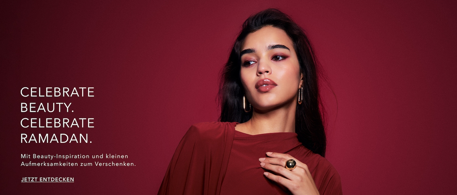 Vor einem weinroten Hintergrund posiert eine Frau mit dunklen, gewellten Haaren, goldenen Ohrringen und einem passenden Ring, während sie ein weinrotes Oberteil trägt; Text sagt: Celebrate Beauty. Celebrate Ramadan. Jetzt entdecken.