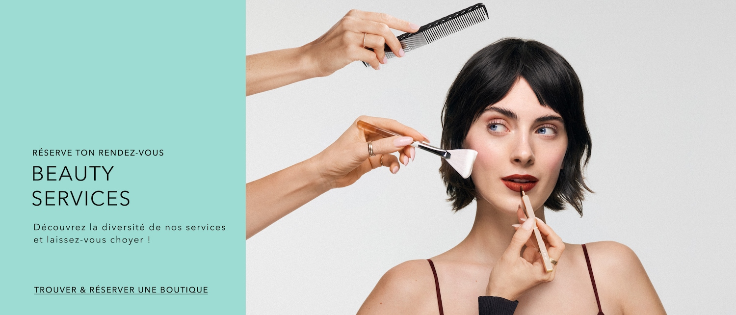 Découvrez nos services de beauté : une femme se fait maquiller les lèvres avec un crayon beige, tandis qu'un pinceau applique du fard sur sa joue et qu'un peigne est tenu au-dessus de sa tête. Réservez votre rendez-vous dès maintenant !