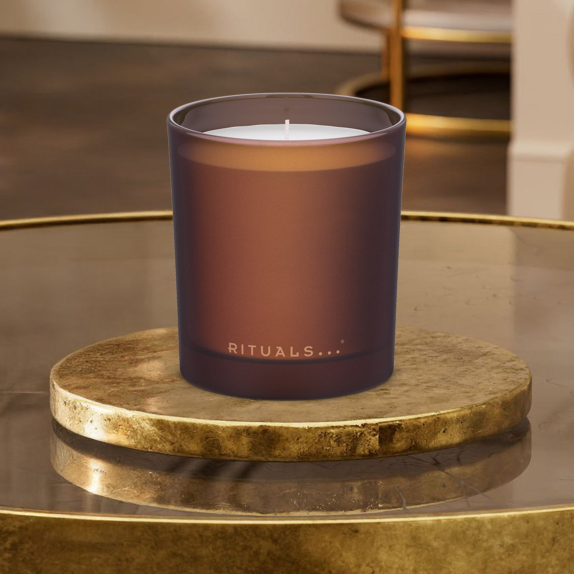 Ein braunes Rituals... Duftkerzenglas sitzt auf einem goldenen Untersetzer und verströmt einen warmen, einladenden Duft.