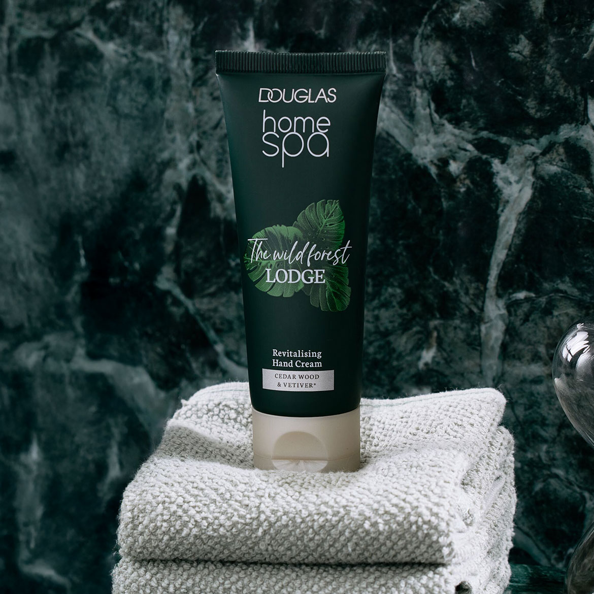 Die Douglas Home Spa Handcreme Wild Forest Lodge in einer dunkelgrünen Tube mit Blättern, die auf einem Stapel grauer Handtücher steht, bietet eine revitalisierende Pflege mit Zedernholz- und Vetiverduft.
