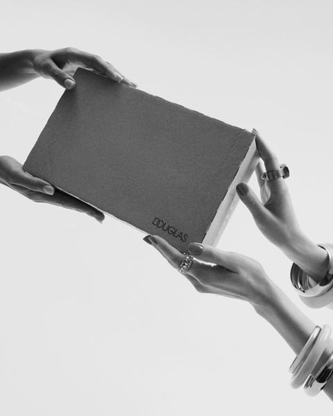 Zwei Hände mit Ringen und Armreifen halten eine graue Douglas-Box, die zum Auspacken bereit ist.