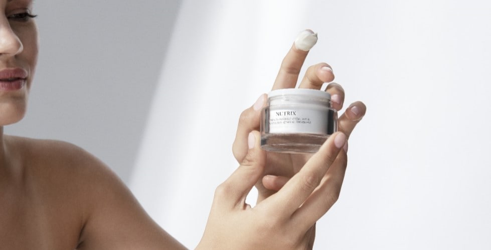 Nahaufnahme einer Frau, die ein Glas Nutrix-Creme mit weissem Deckel hält und eine kleine Menge Creme auf ihren Finger nimmt.