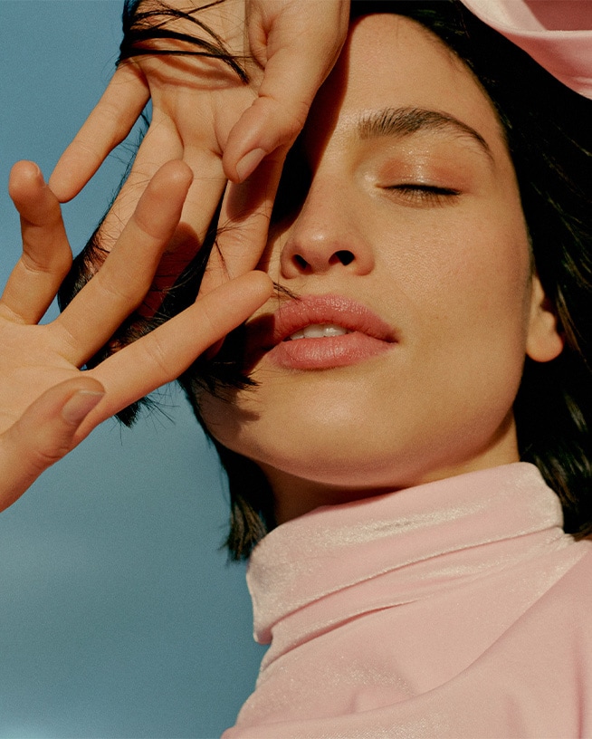 Gros plan sur un visage féminin avec les yeux fermés et les mains délicatement placées, peau éclatante et lèvres roses, le tout sur un fond bleu doux.